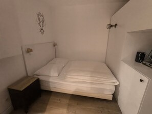 A small, single-bed room with a wooden nightstand, a wall-mounted shelf, and a bed with white bedding.