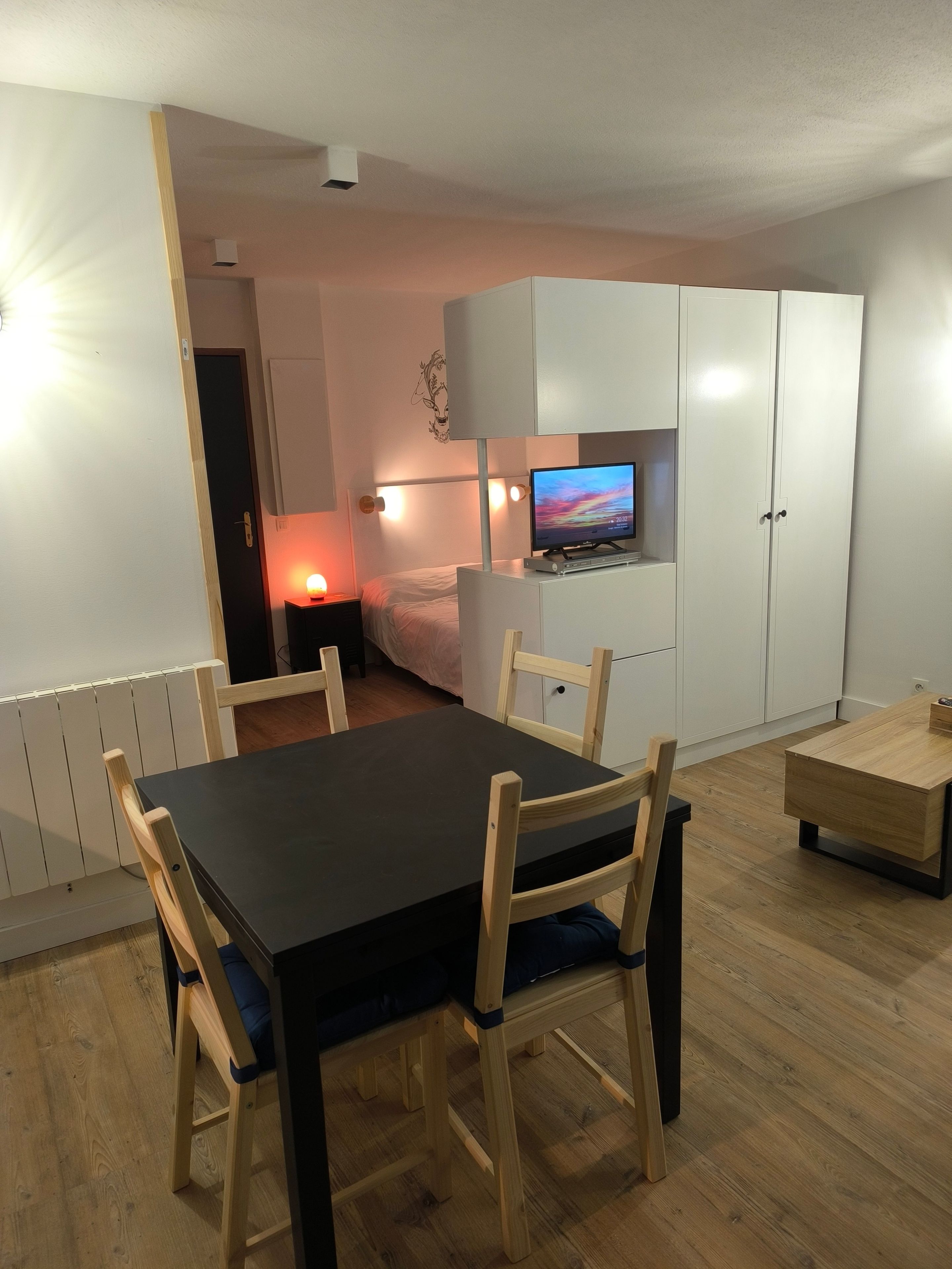 A room with a dining table, chairs, a bed, a TV, and a wardrobe.