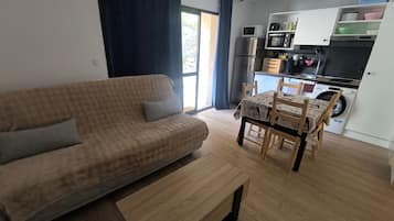 A living room with a sofa, a dining table with chairs, a kitchen area with appliances, and a large window with curtains.