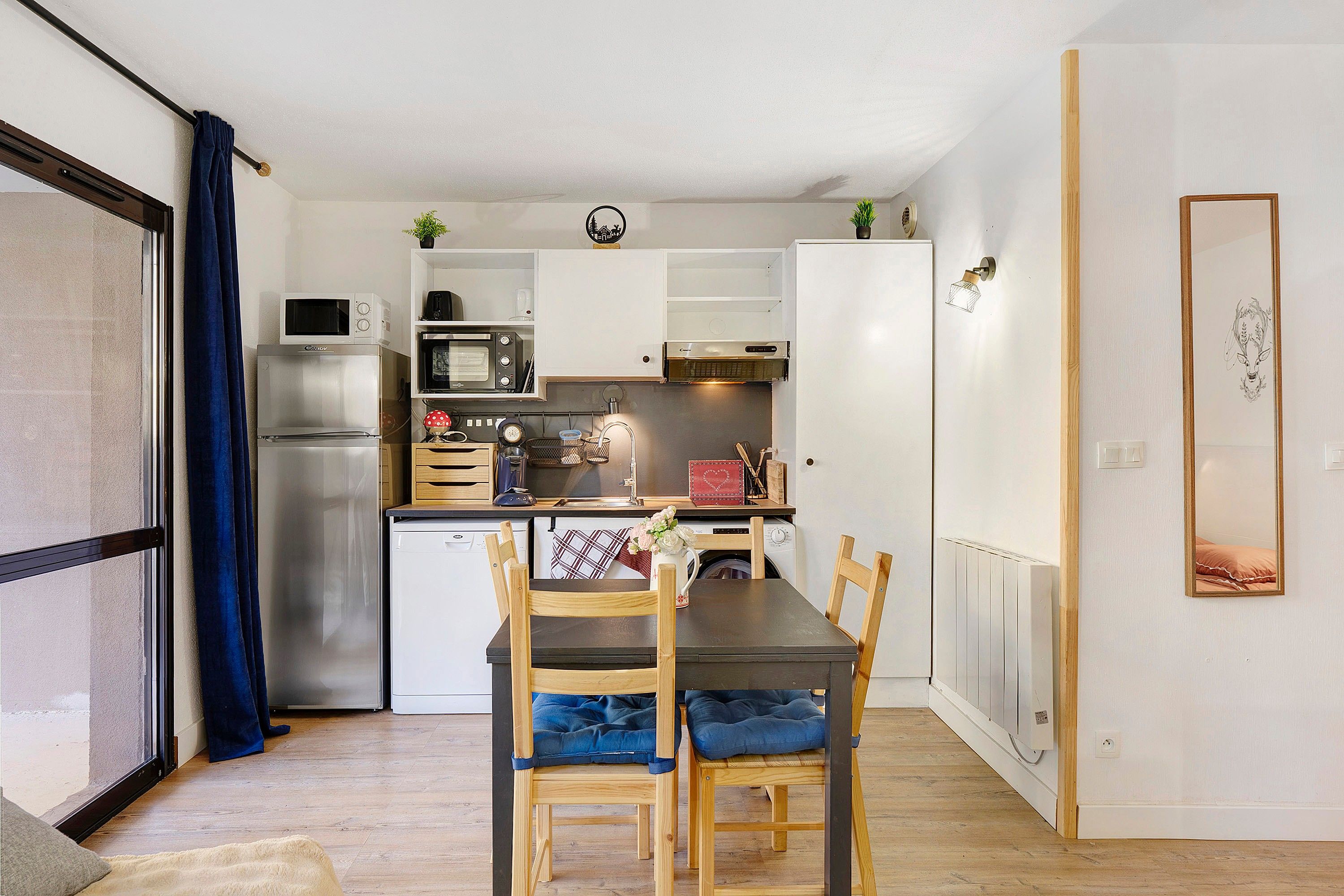 A compact kitchen with a refrigerator, microwave, and stove, a dining table with chairs, and a small window with curtains.