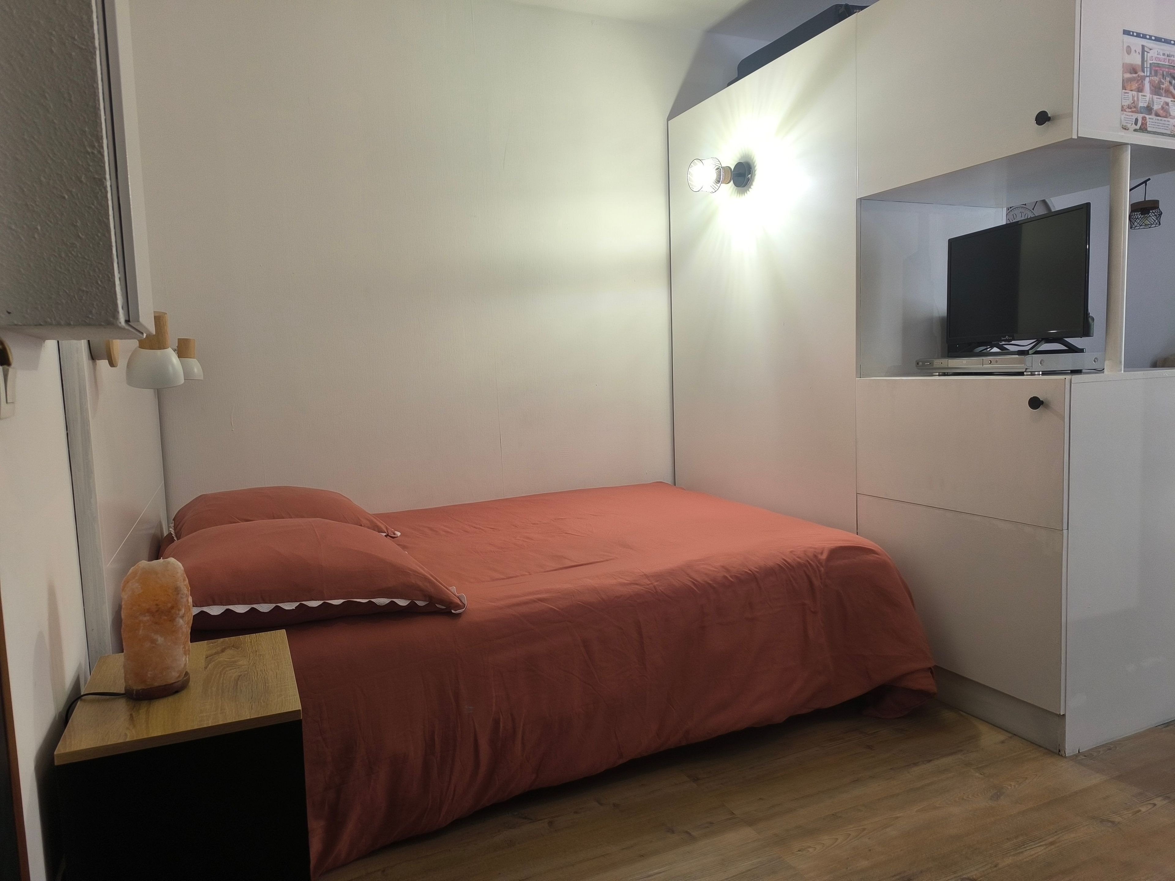 Una camera da letto con un letto, un comodino, un termosifone, una televisione su un mobile e una lampada a parete.