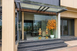 A modern building entrance with glass doors, a small outdoor seating area, and a large potted plant with orange flowers.