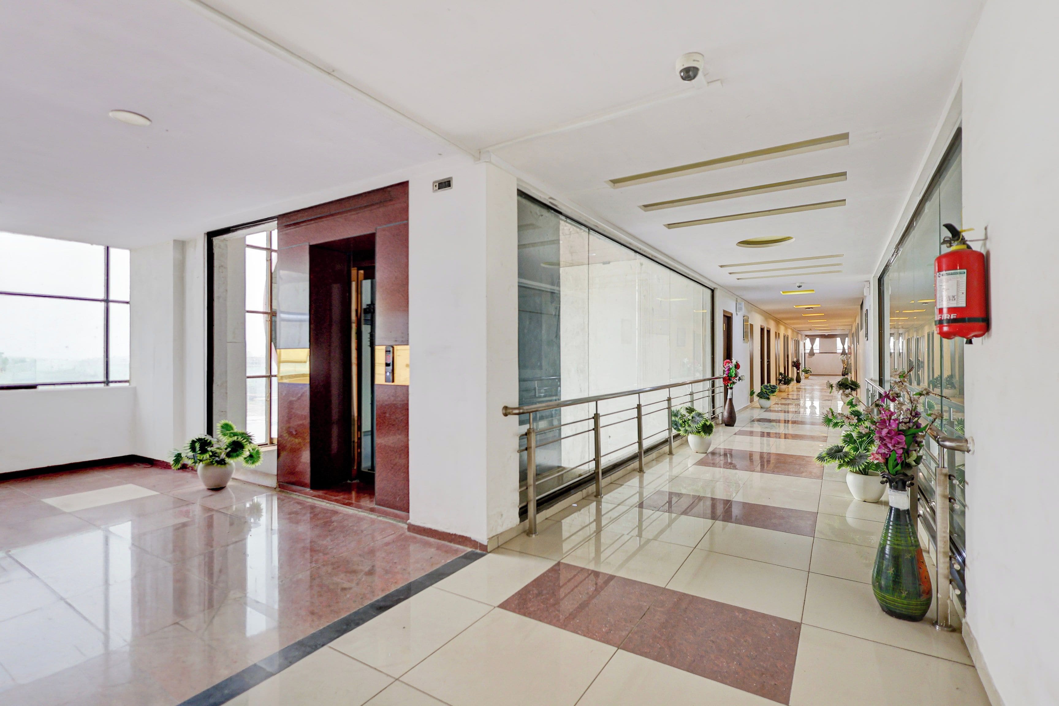A spacious corridor with a fire extinguisher mounted on the wall, a glass door, and potted plants along the walkway.