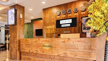 Un mostrador de recepción con detalles de madera, un cartel que indica 'Reception' y 'Palette' con horarios de registro de entrada y salida, y relojes que muestran diferentes zonas horarias.