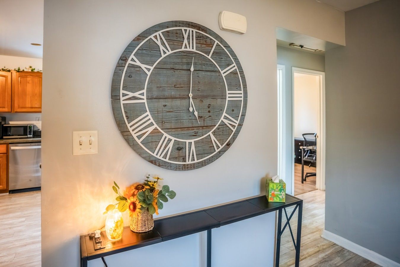 Une horloge murale avec des chiffres romains, une petite table sur laquelle est posé un vase de fleurs et une boîte à mouchoirs.