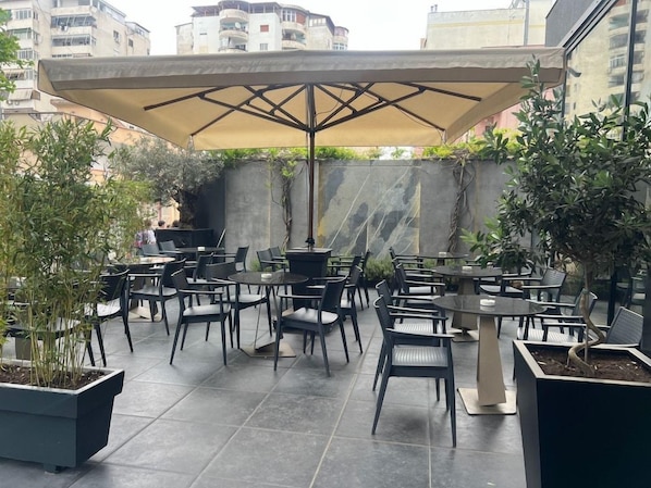 An outdoor patio area with multiple tables and chairs, some covered by umbrellas, and potted plants. - Moncafe Hotel Boutique Spa (Tirana)