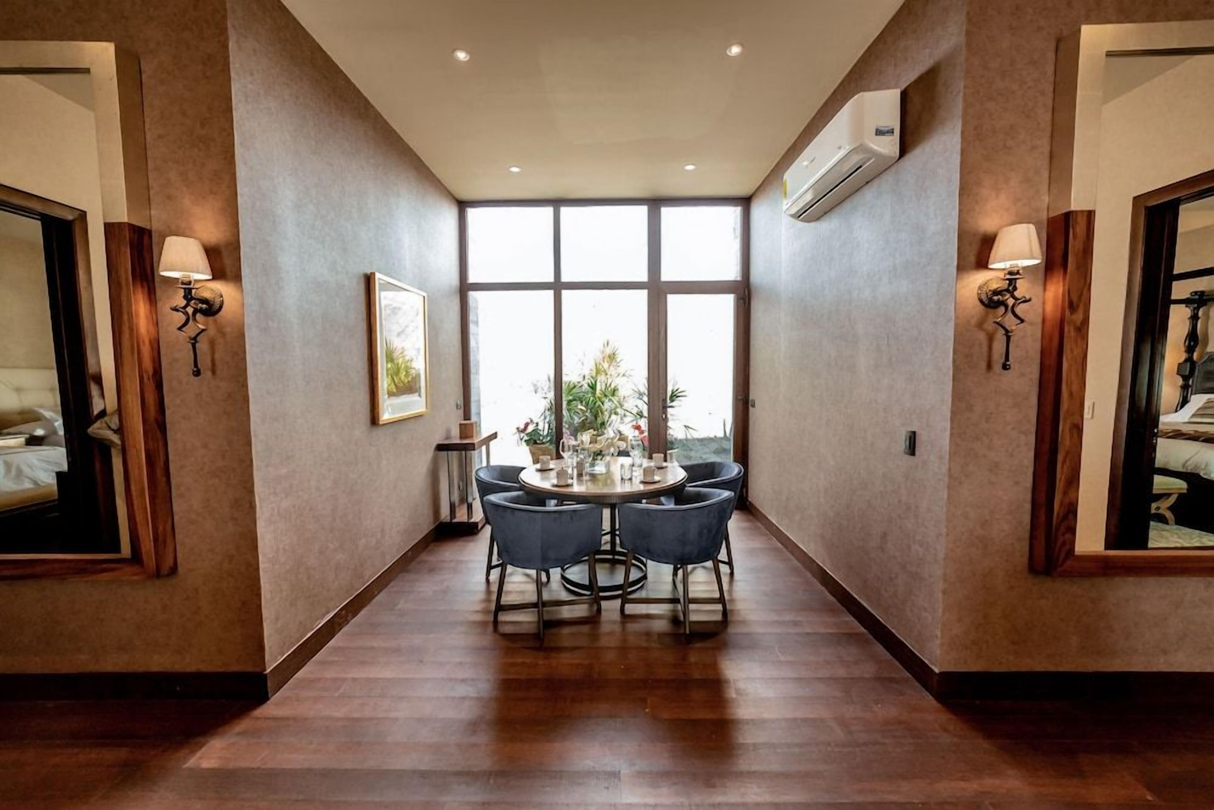 A hotel room with a large window, a round dining table with chairs, and two wall-mounted lamps.