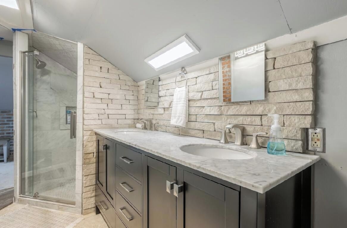 Une salle de bain dotée d’une douche à parois de verre, d’un meuble-lavabo à deux vasques et d’un mur arrière en pierre.