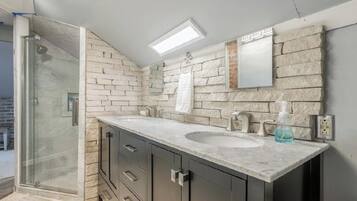 Une salle de bain dotée d’une douche à parois de verre, d’un meuble-lavabo à deux vasques et d’un mur arrière en pierre.