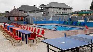 Piscina com deck azul, mesa de pingue-pongue e fileiras de cadeiras.