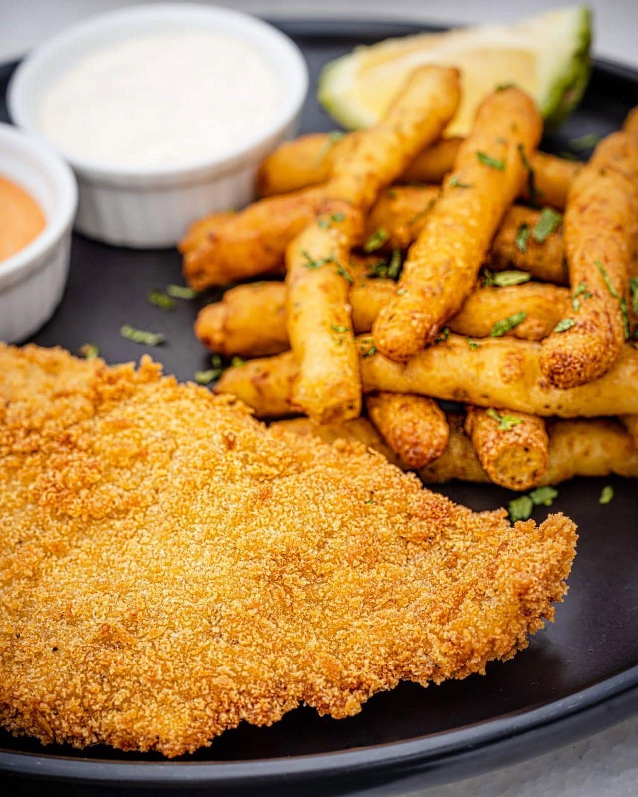 A plate of breaded and fried food items, possibly chicken tenders, with a breaded cutlet and a side of dipping sauce.