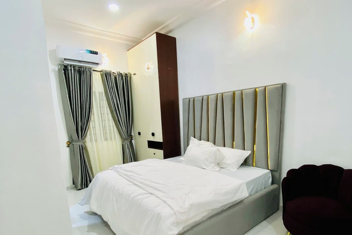 A modern bedroom with a large bed, a dark-colored armchair, and a wall-mounted air conditioning unit.