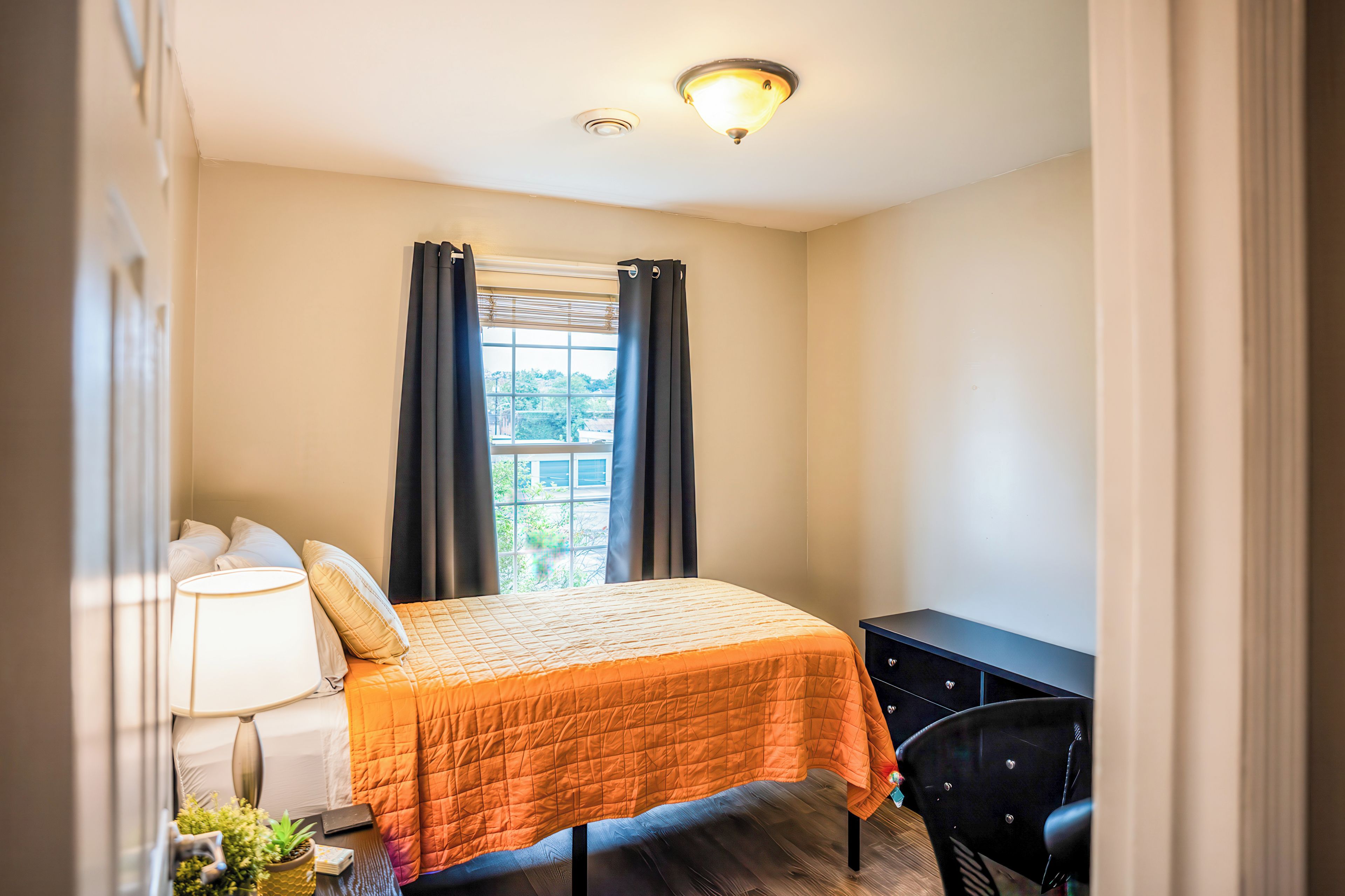 Une chambre à coucher avec un lit, un bureau, une chaise, une lampe et une fenêtre avec des rideaux.