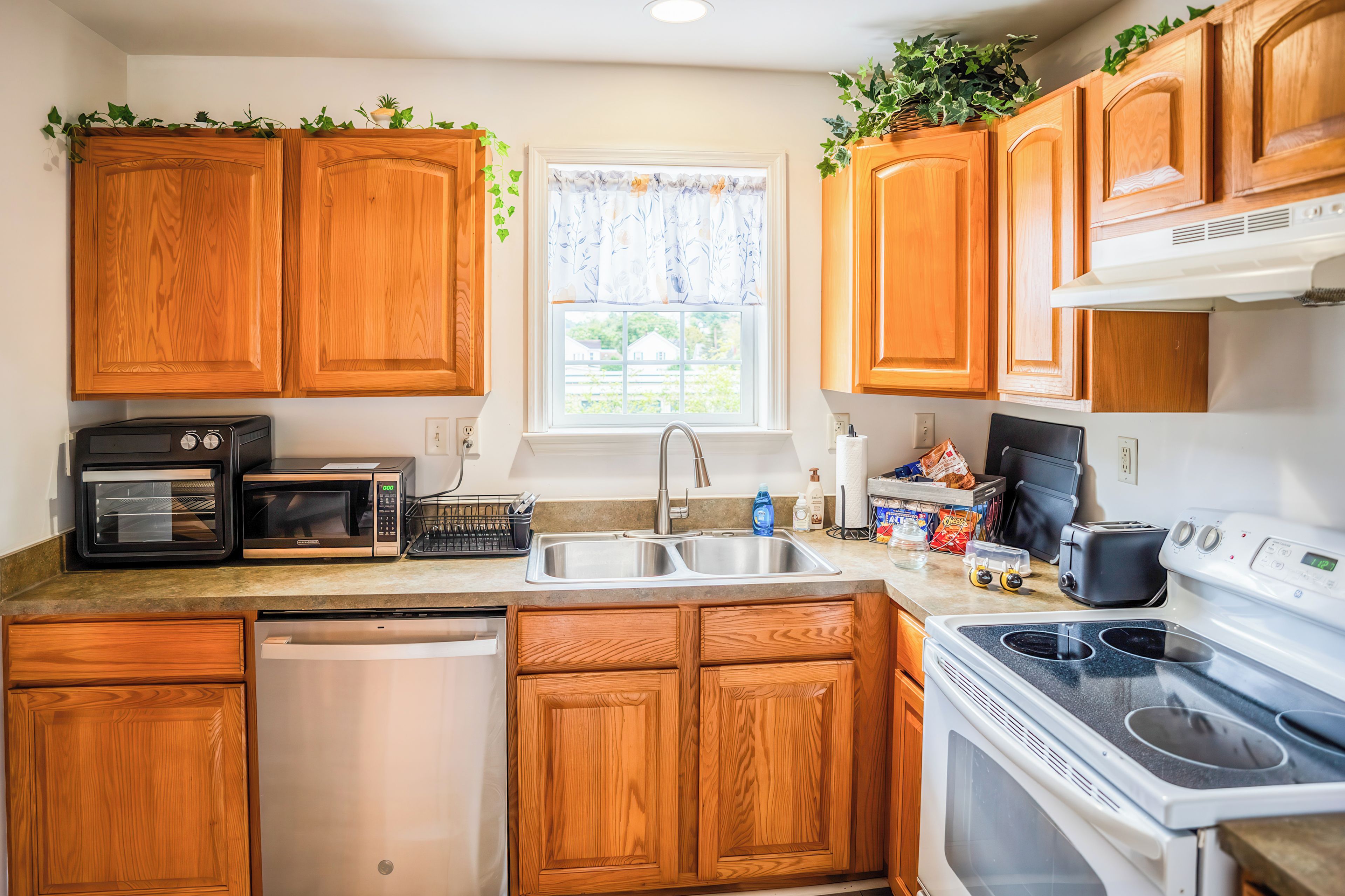 A kitchen with wooden cabinets, a dishwasher, a microwave, a stove, and a window with a view of houses.