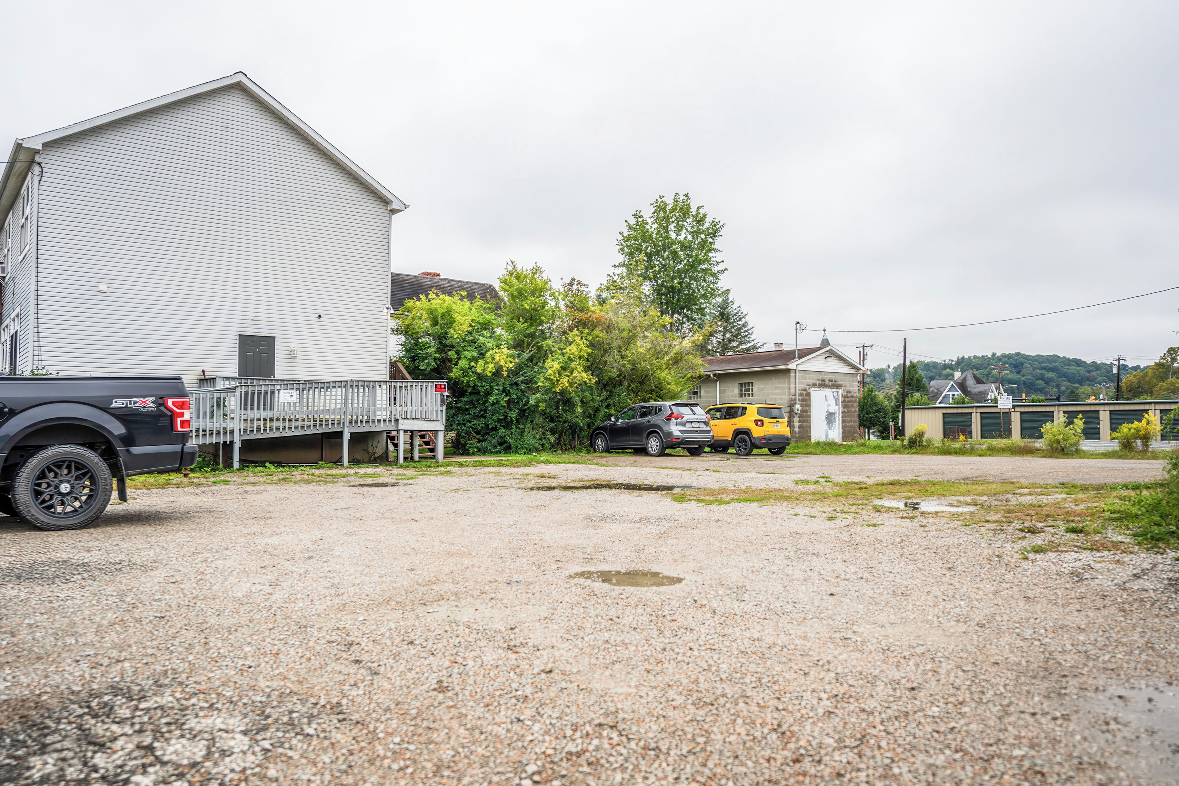 A gravel parking area with a large white building and a smaller structure in the background.