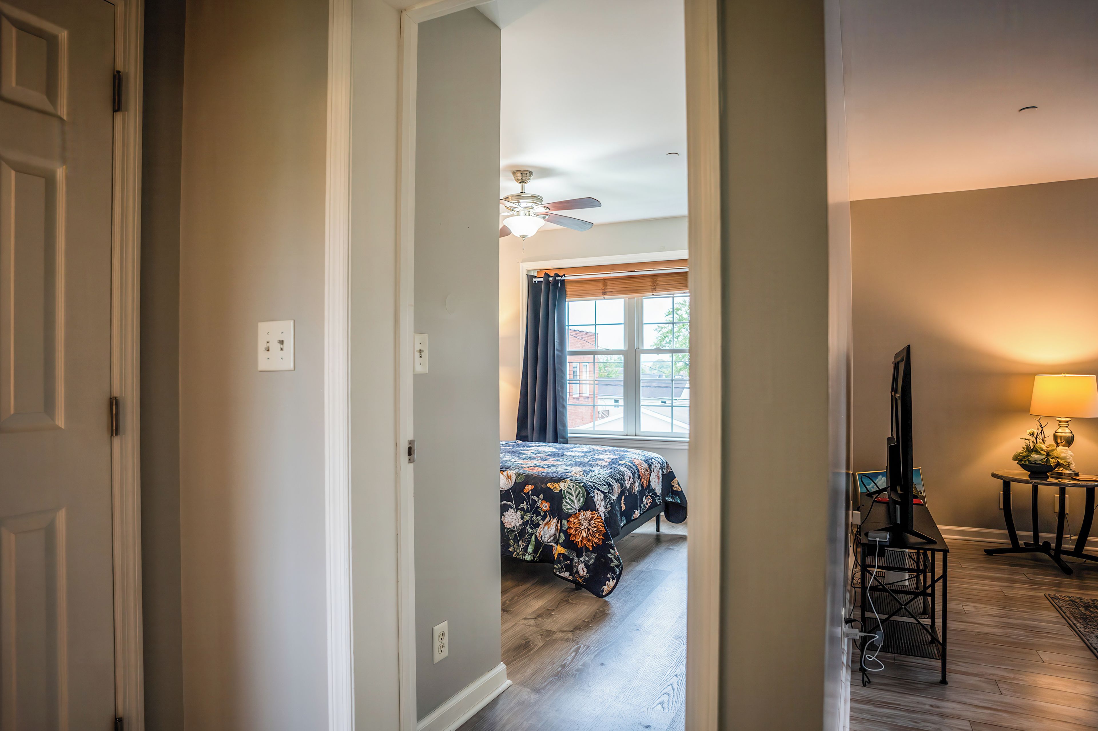 A hallway leading to a bedroom with a bed, a ceiling fan, and a window with curtains.