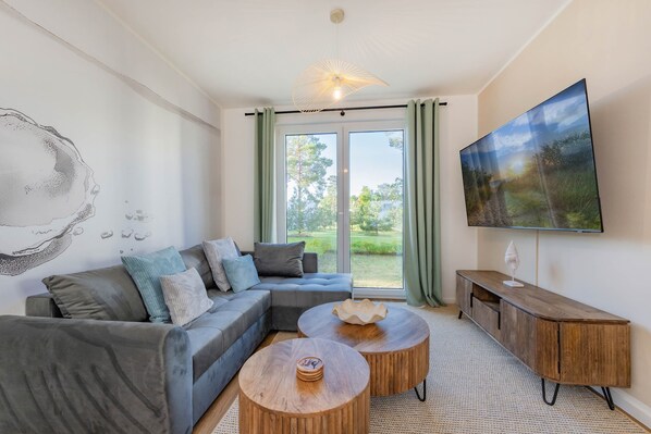 A modern living room with a sectional sofa, two round wooden coffee tables, a flat-screen TV, and a large window with a view of greenery. - Modern vacation rental with a terrace, sauna, and recreation area (Zirchow)
