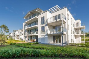 Um edifício residencial moderno com vários andares, varandas, telhado plano e um jardim bem cuidado.