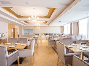 A hotel dining area with multiple tables and chairs, a central chandelier, and a kitchen in the background. - Vienna Hotel (Chengdu)