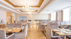 A hotel dining area with multiple tables and chairs, a central chandelier, and a kitchen in the background. - Vienna Hotel (Chengdu)