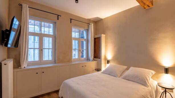 A bedroom with a bed, two windows with curtains, a wall-mounted TV, a bedside lamp, and a radiator. - Le Sablon Retreat Private En Suite Room (Brussels)