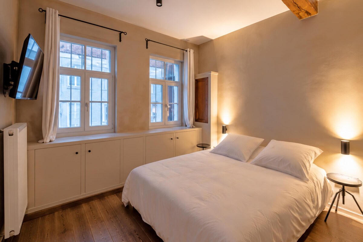 A bedroom with a bed, two windows with curtains, a wall-mounted TV, a bedside lamp, and a radiator.