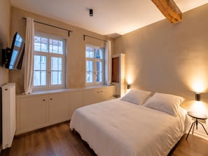 Un dormitorio con cama, dos ventanas con cortinas, televisor montado en la pared, lámpara de noche y radiador.