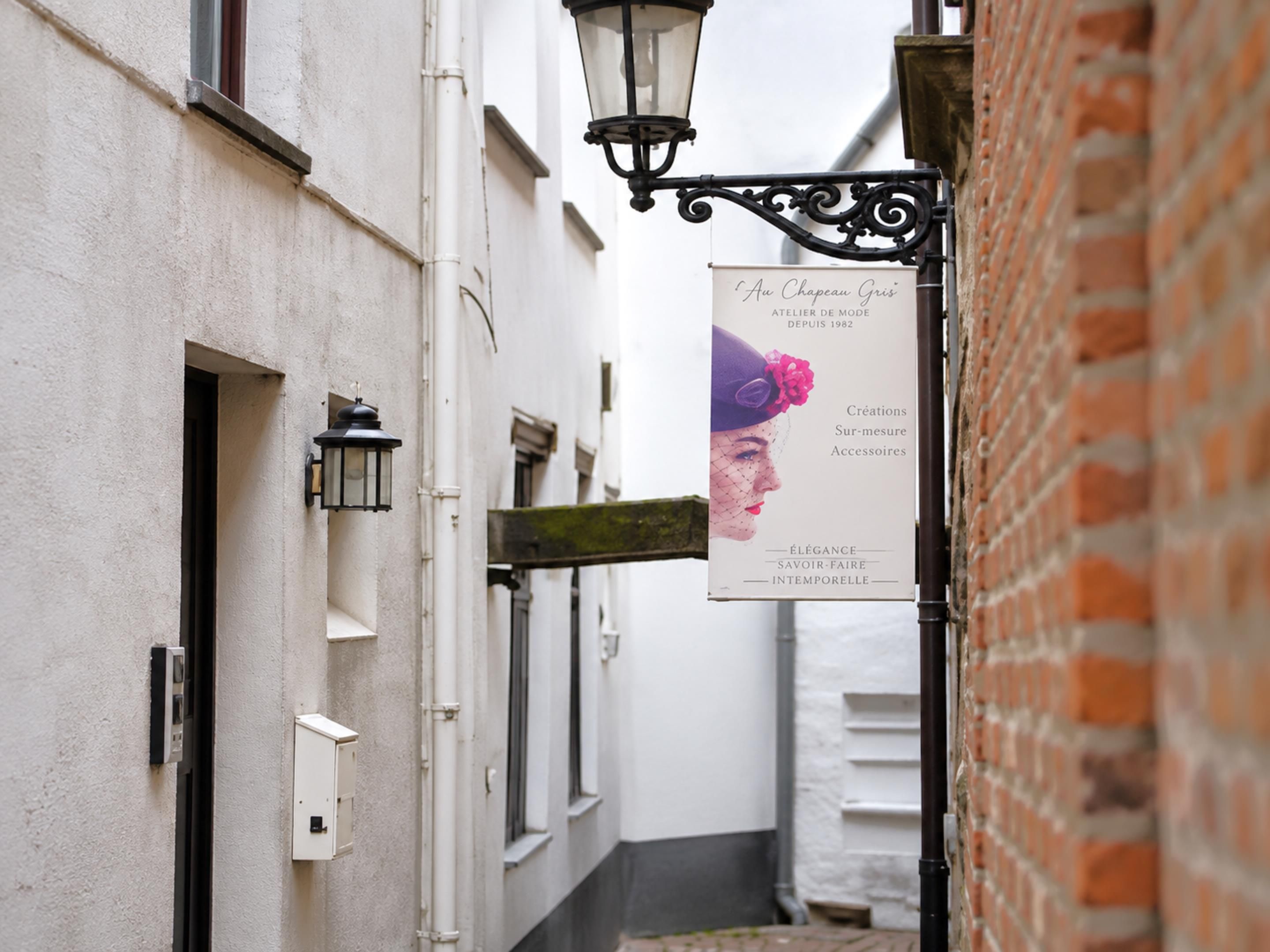A narrow alley with a sign for 'Au Chateau Gris' boutique, a white building with a brick wall, and a hanging street lamp.