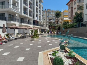 A poolside area with lounge chairs and umbrellas, surrounded by multi-story buildings with balconies. - Qoople Elize C15 - Cozy 1-bedroom apartment in Alanya (Alanya)