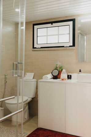 A bathroom with a glass shower enclosure, a toilet, a sink with a mirror, and a window above the sink. - Casa da Venda by Lethes Legacy (Refóios do Lima)