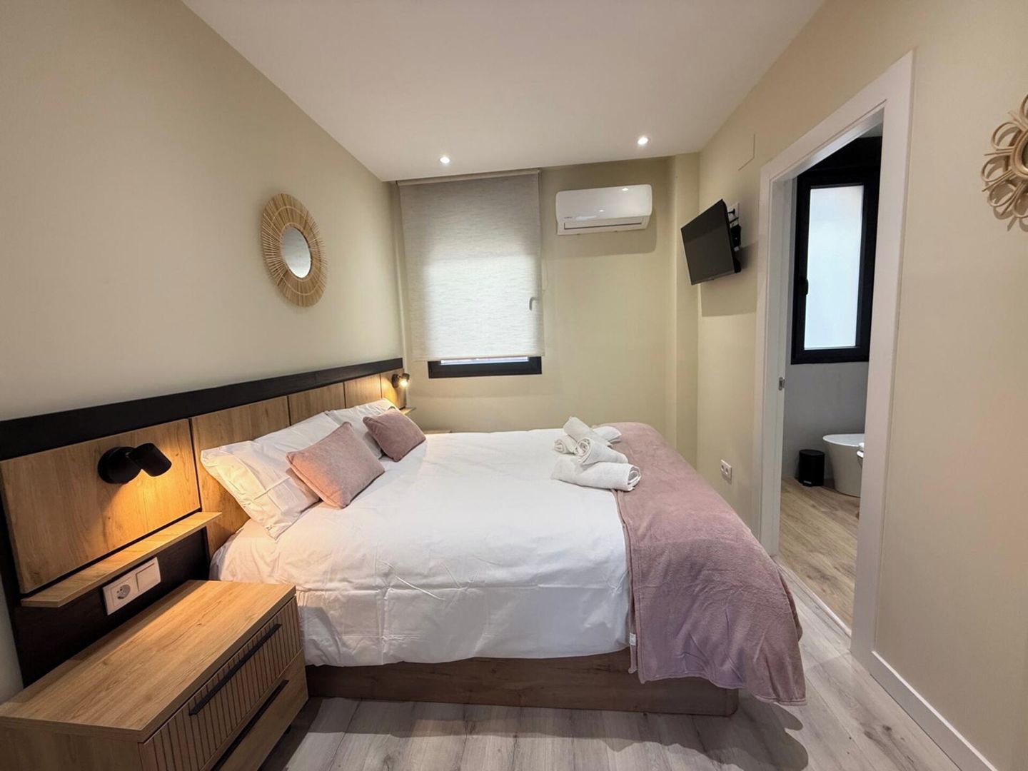 Una habitación de hotel con cama, mesitas de noche, televisor montado en la pared y ventana con persianas.