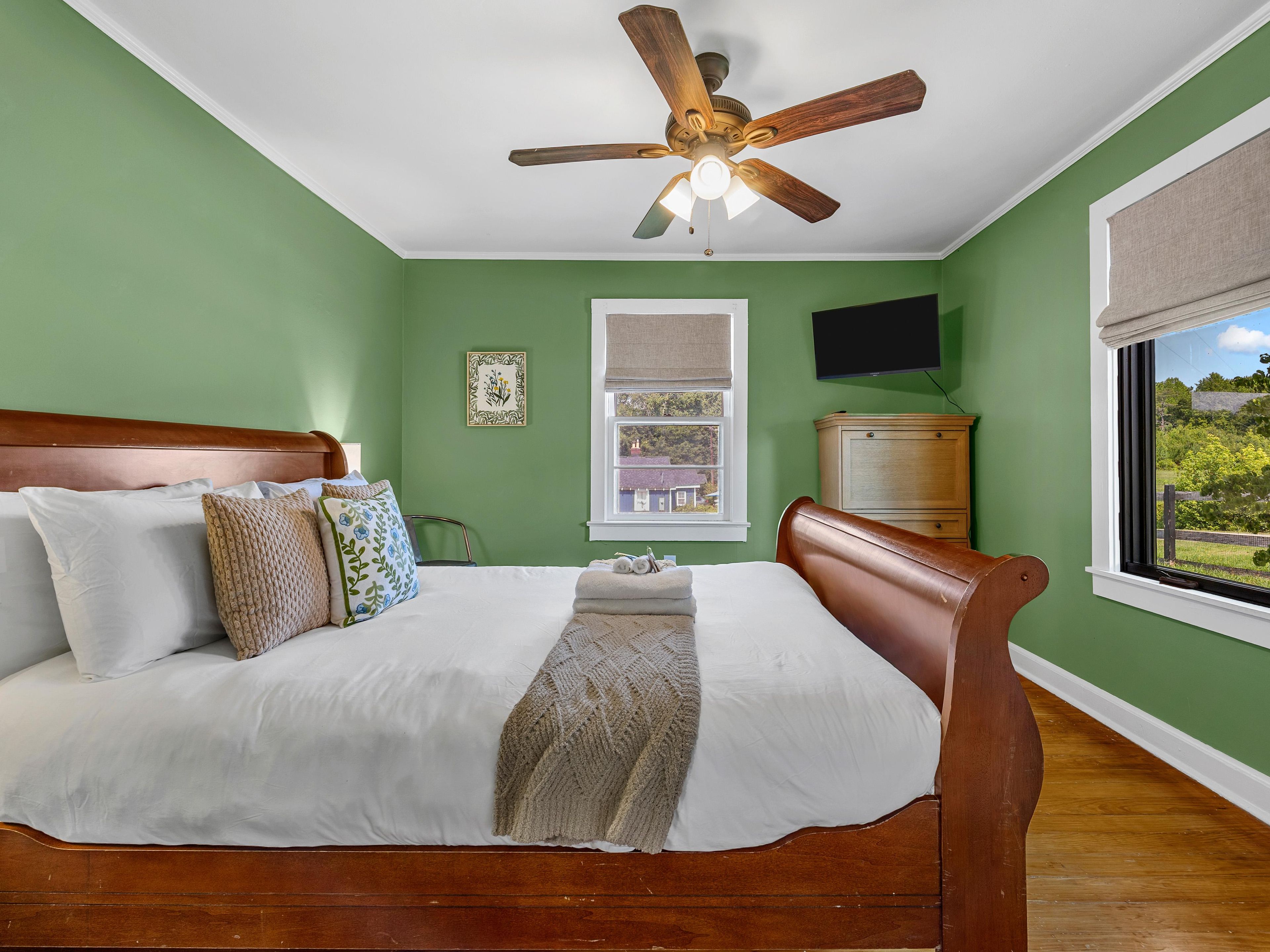 A bedroom with a wooden bed, a ceiling fan, a window with a view, and a television on the wall.