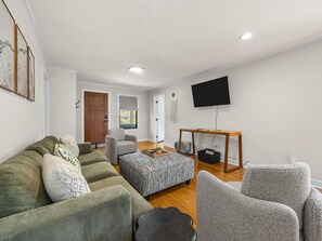 Una sala de estar con sofá, sillón, reposapiés y televisión montada en la pared.