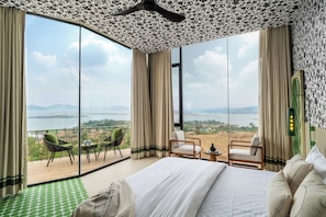 A hotel room with a large bed, two chairs, a small table, and a view of a lake and mountains.