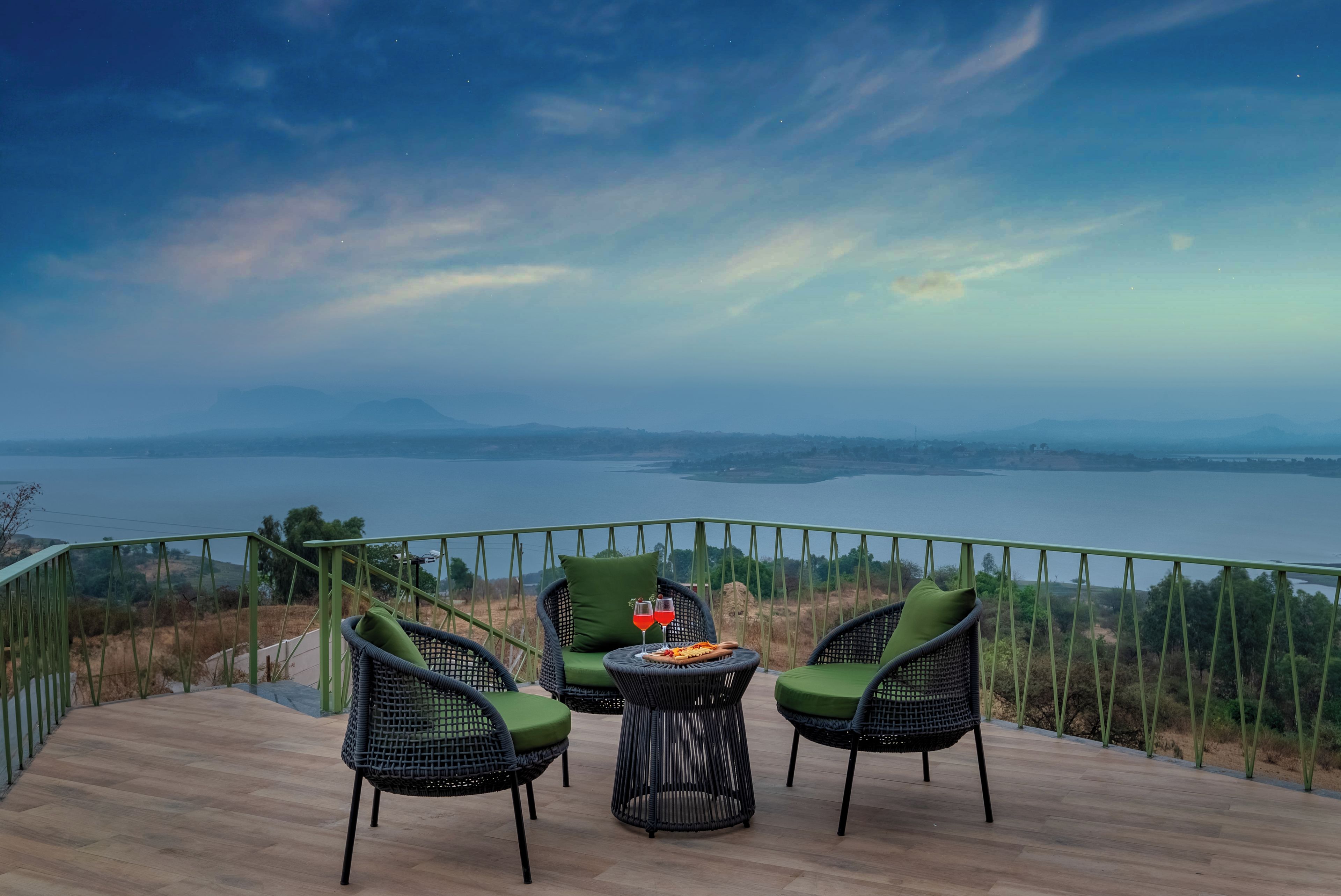 Una terraza con sillas de mimbre y una mesa, con vistas a un cuerpo de agua y montañas distantes.