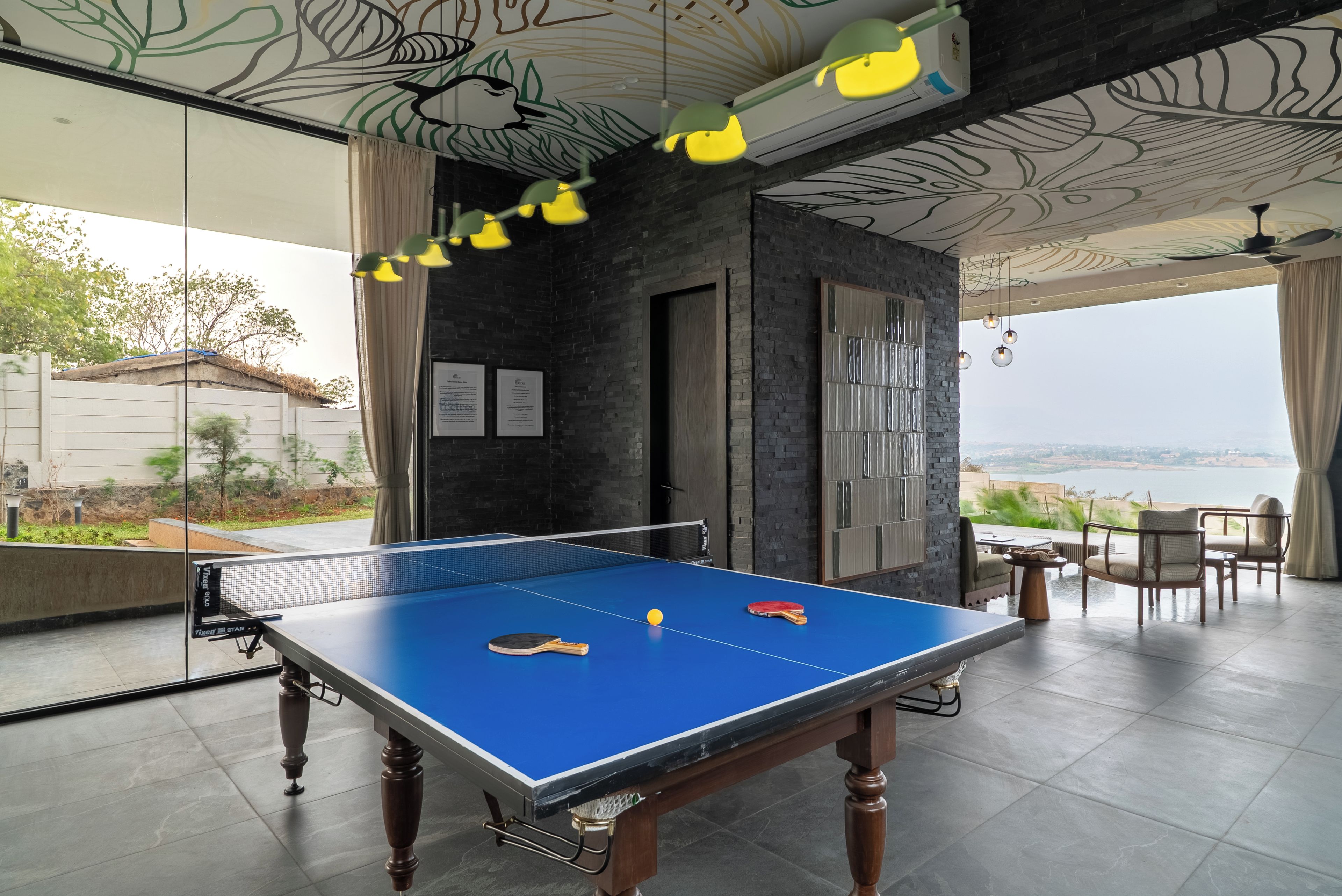 A ping pong table with two paddles and a ball, a ceiling with leaf patterns, and a view of a body of water.