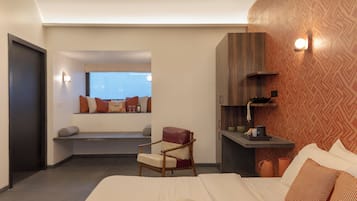 Habitación de hotel con cama, sillón de lectura, un pequeño rincón de estar con ventana y estante empotrado con diversos objetos.