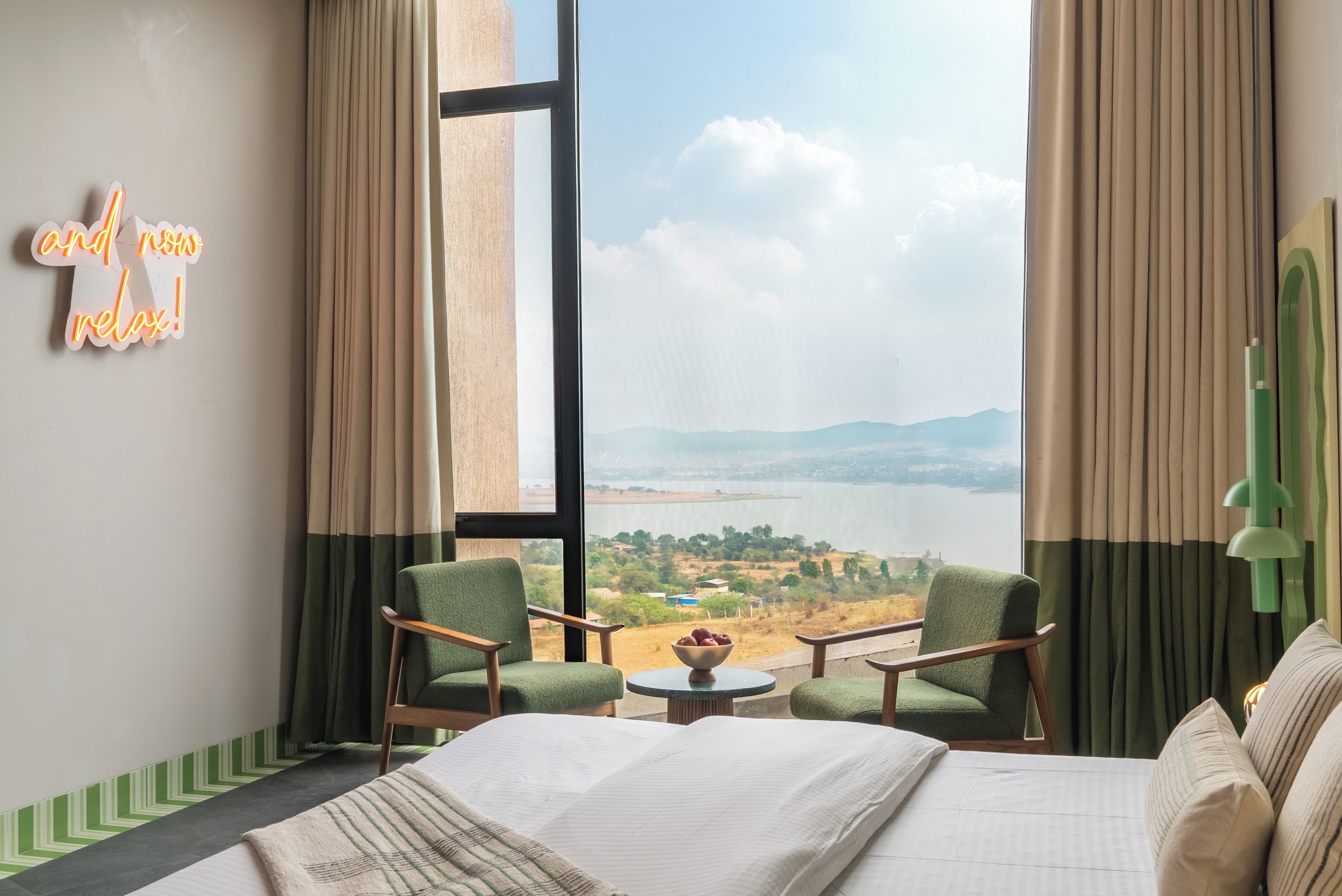 Habitación de hotel con cama, dos sillas, una mesita con frutero, y vistas a un lago y montañas a través de amplios ventanales.