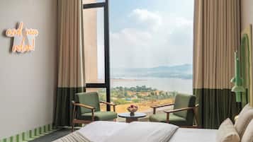 Habitación de hotel con cama, dos sillas, una mesita con frutero, y vistas a un lago y montañas a través de amplios ventanales.