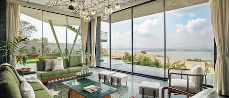 Un salón amplio con un sofá verde, una mesa de centro y vistas a una playa y al océano a través de ventanas grandes.