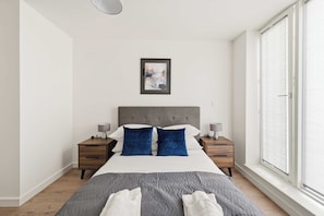 A modern bedroom with a large bed, two nightstands, a grey headboard, a framed picture on the wall, and a window with blinds. - Modern & Bright 1BED Central Birmingham Apartment (Birmingham)