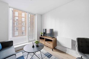 Una sala de estar moderna con una televisión de pantalla plana sobre un mueble de madera, un sofá gris, una pequeña mesa redonda de centro y una planta en maceta.