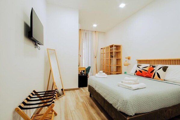 A hotel room with a bed, a TV mounted on the wall, a wooden chair, a small table, and a bookshelf. - Lara's Home (Naples)