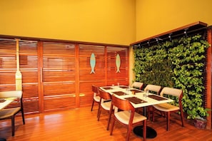 Un comedor con mesa y sillas de madera, una pared con persianas verticales y una pared cubierta de plantas verdes.