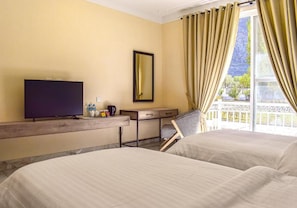 A hotel room with two beds, a television, a desk with a chair, a window with curtains, and a view of greenery outside.