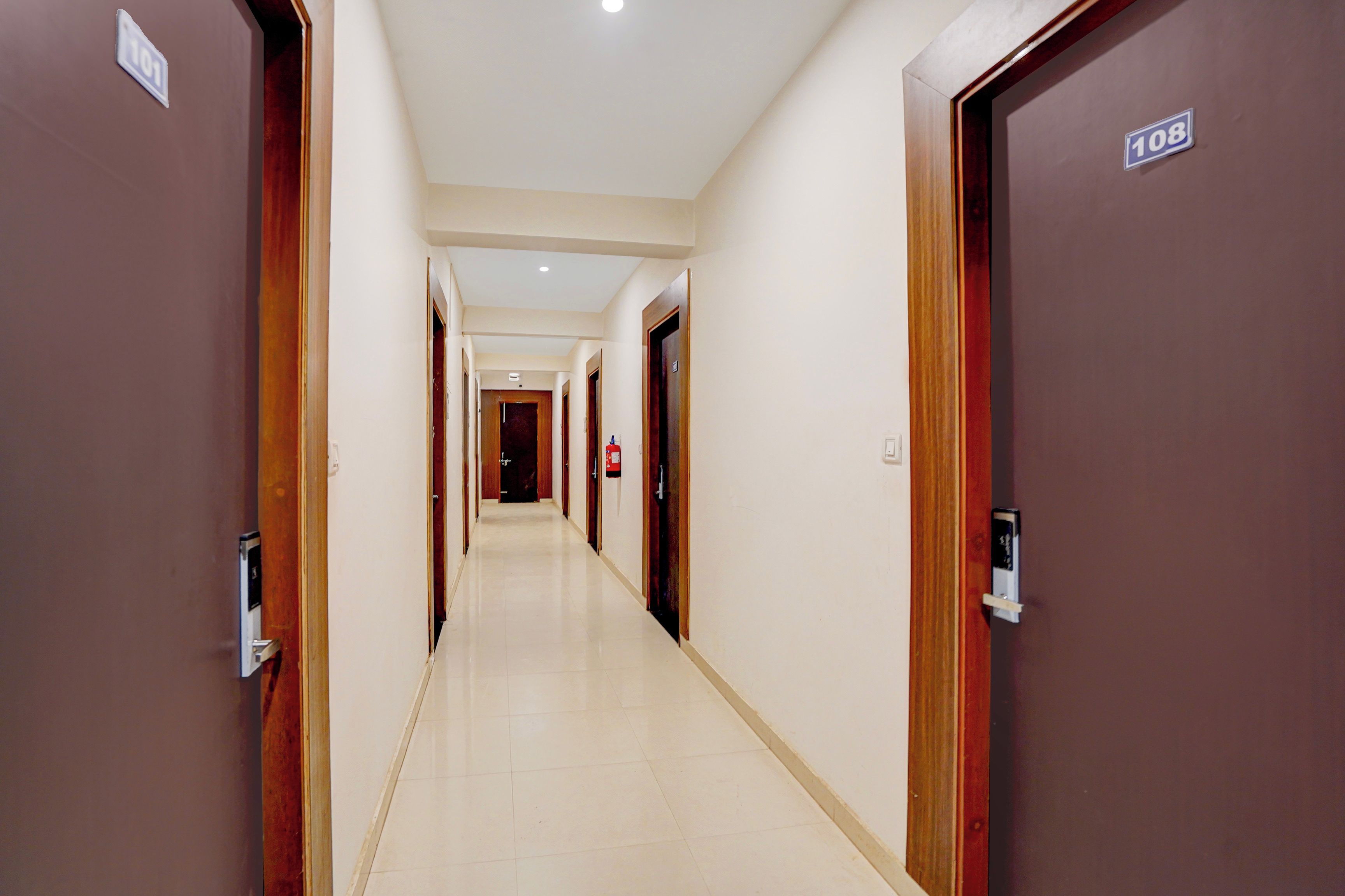 Un corridoio d'albergo con porte provviste di numeri civici, un estintore e un lampadario a soffitto.