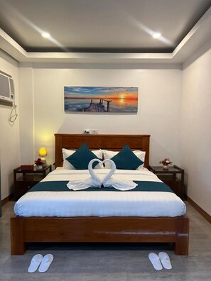 A hotel room with a wooden bed, white bedding, teal pillows, a painting of a sunset, a nightstand with a lamp, and a vase with flowers. - Ocean Star Hotel Coron (Coron)