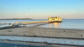 A pier extending into calm waters with a building at the end, a sandy beach in the foreground, and a clear sky.