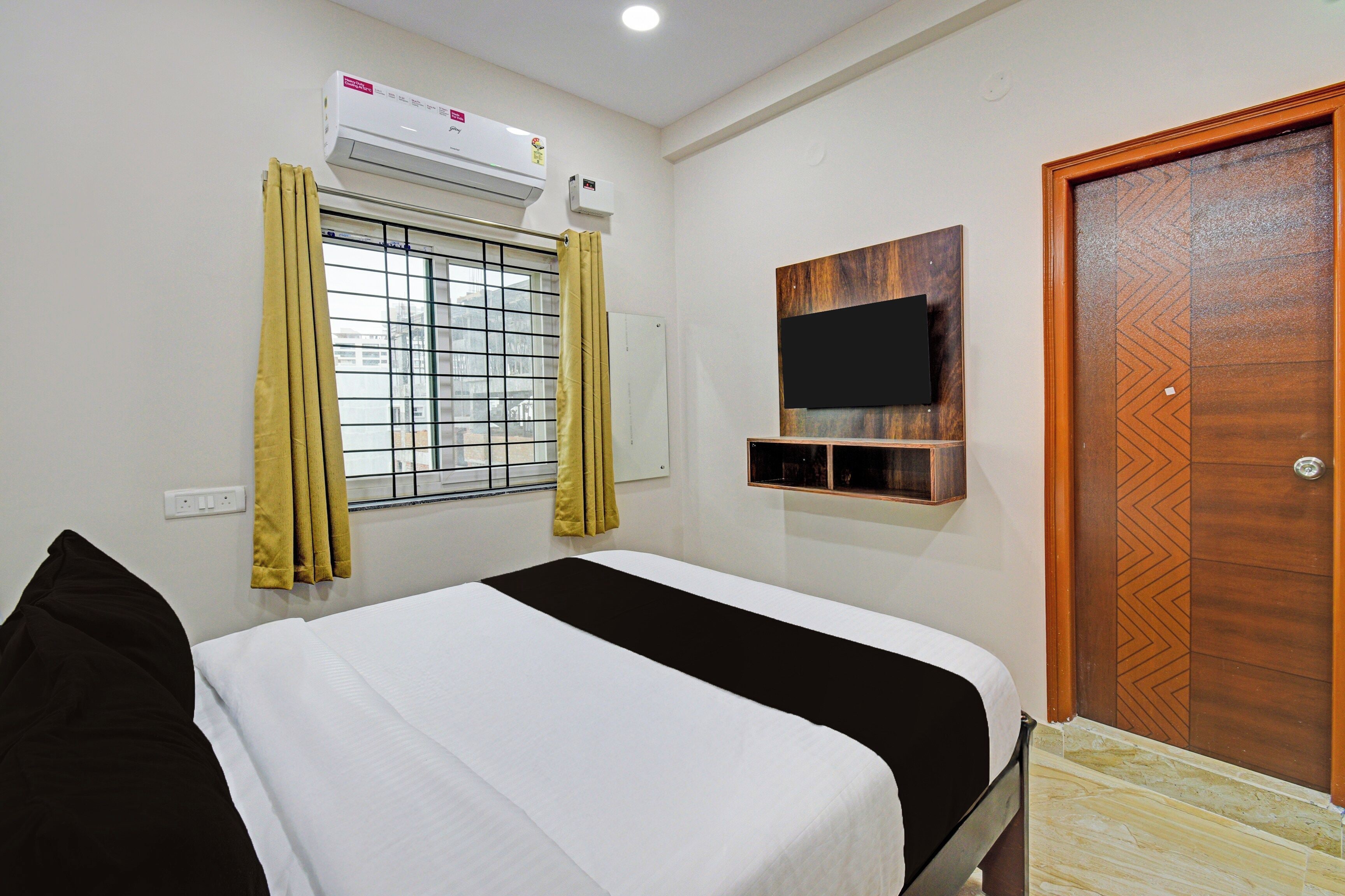 Una habitación de hotel con una cama, un equipo de aire acondicionado, una ventana con cortinas, un televisor montado en la pared y una puerta de madera.