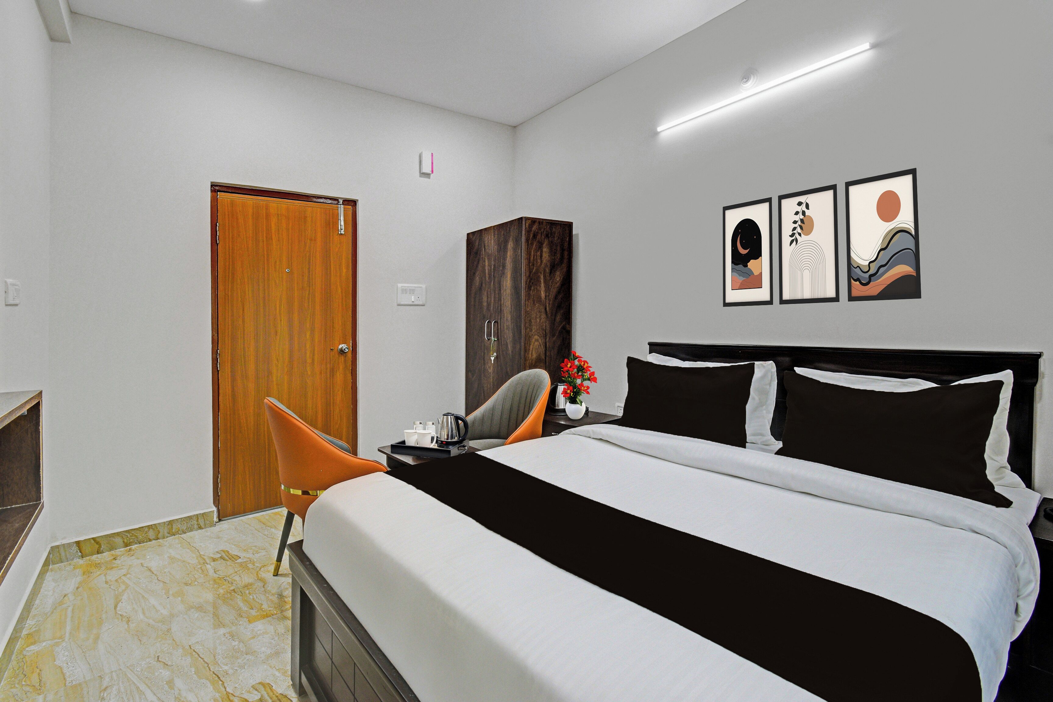 A hotel room with a bed, a wooden wardrobe, a desk with a chair, and artwork on the wall.